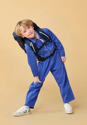 Blauer Hoodie-Overall mit langen Ärmeln, kombiniert mit einem schwarzen Rucksack. Die Schuhe sind weiße Sneakers mit einem grünen Logo. Der Hintergrund ist hellbeige.