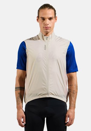 Leichte, beige Fahrradswesten mit hohem Kragen, Reißverschluss vorne und elastischem Saum. Getragen über einem blauen Kurzarmshirt.
