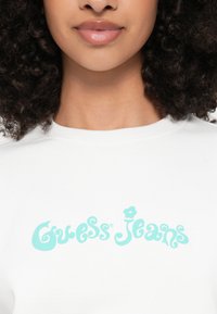 Λευκό βαμβακερό μπλουζάκι που φέρει το λογότυπο "Guess Jeans" σε μέντα πράσινο χρώμα σε παιχνιδιάρικη γραμματοσειρά, με ριμπ λαιμόκοψη και κοντά μανίκια.