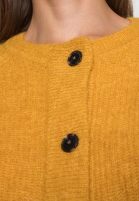 Senapsgul, stickad cardigan med rund halsringning, försedd med två stora svarta knappar och ett texturerat, mjukt tyg.