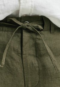 Olivgrüne Leinenhose mit einer vorn gebundenen Schleife, breitem Bund und seitlichen Taschen, kombiniert mit einem weißen Hemd mit Knöpfen.