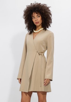 Vrouw met krullend haar, gekleed in een beige wikkeljurk met lange mouwen, vastgemaakt met een decoratieve speld en een grote witte kralenhalsketting.