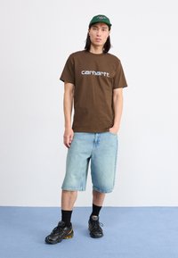 Hnedé bavlnené tričko s bielym logom "carhartt.", skombinované s svetlo modrými džínsovými šortkami. Čierne topánky s žltými akcentmi a čierne ponožky.