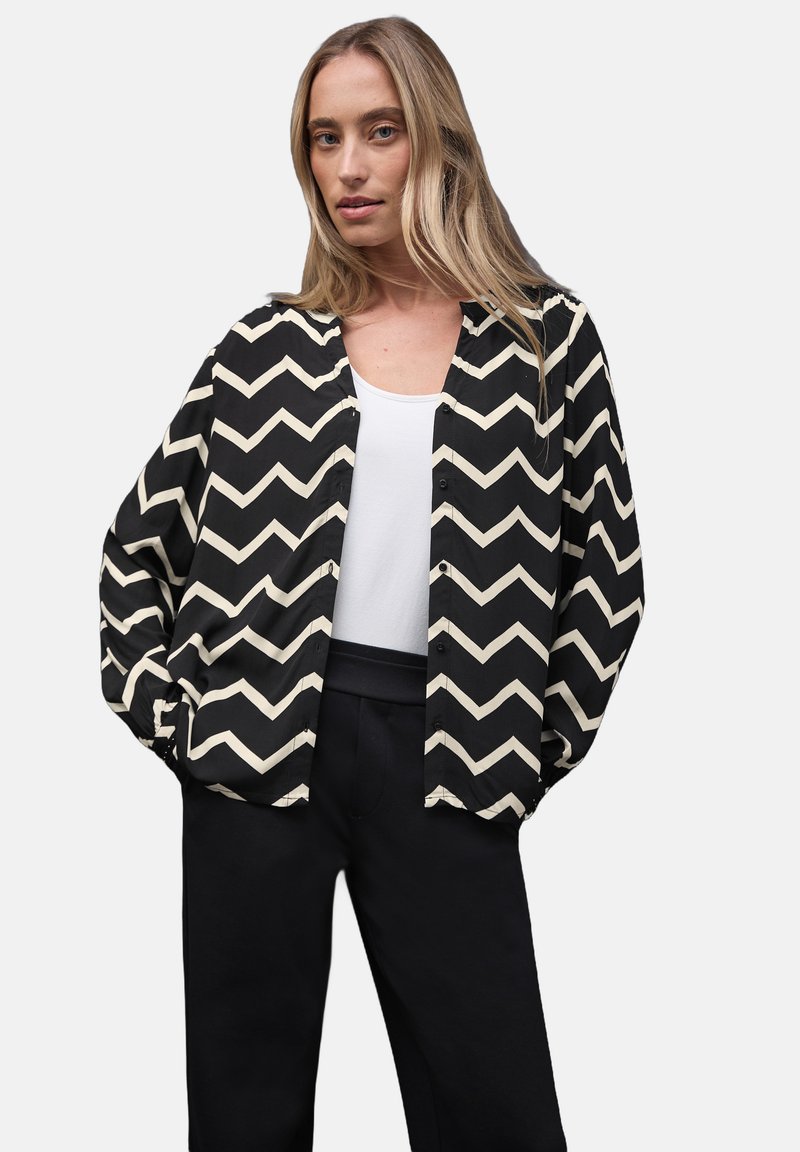 Schwarz-beige, im Zickzackmuster gestaltete Hemd mit Knöpfen, langen Ärmeln, lässiger Passform und nahtlosem Design. Kombiniert mit dunkler Hose.