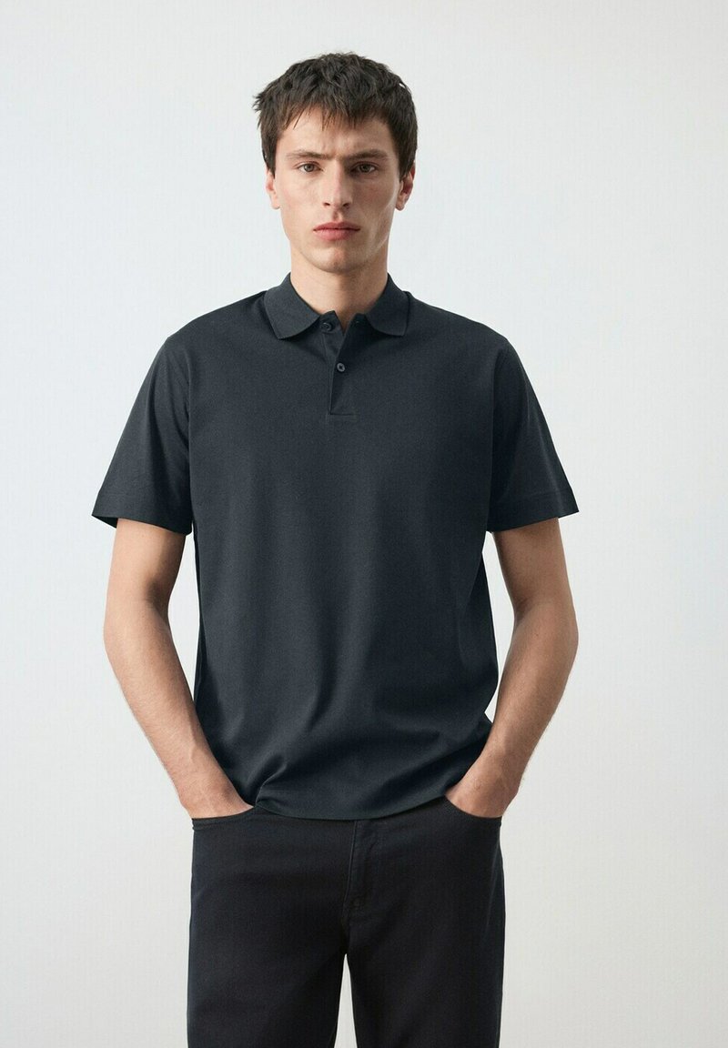 Giovane uomo con capelli corti e scuri, indossa una polo nera a maniche corte e pantaloni neri, in piedi con le mani nelle tasche.