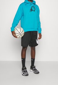 Felpa teal con grafica nera di un pallone da basket, pantaloni corti neri, calzini neri Nike e sneaker grigie/neri. Tiene in mano un pallone da basket bianco.