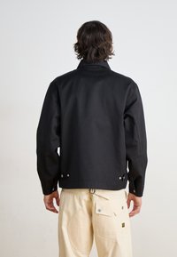Veste en coton noire avec un col montant, des manches longues et des détails de bouton-pression à la taille. Portée avec un pantalon couleur crème.