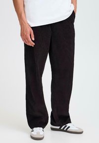 Schwarze Cordhosen mit weitem Bein, die vertikale Rippung und seitliche Taschen aufweisen, kombiniert mit weißen und grauen Turnschuhen.