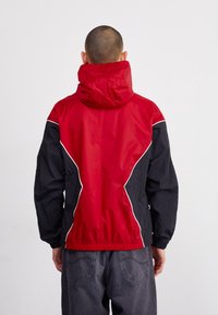 Červeno-černá větrovka s kapucí, bílé lemování a volný střih. Texturovaná látka, elastické manžety a lem.