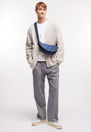 Joven con cárdigan beige, camisa blanca, pantalones grises, zapatillas blancas y un bolso cruzado azul marino, de pie con las manos en los bolsillos.