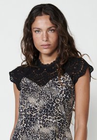 Top senza maniche con colletto in pizzo nero caratterizzato da motivi intricati e un corpo in tessuto beige con design a stampa leopardata in toni scuri.