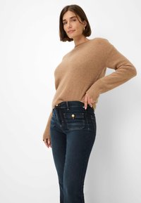 Beige gestrickter Pullover mit langen Ärmeln und lässiger Passform, kombiniert mit dunkelblauen Jeans mit Taschen vorne und goldfarbenen Hardware-Details.