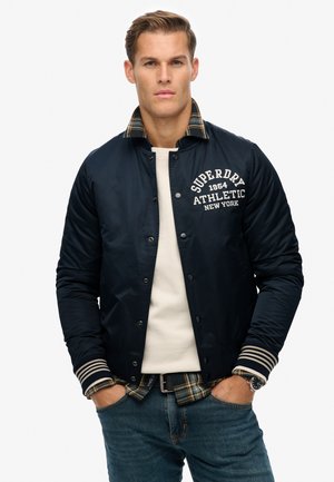 Navy blue bomber jacket with quilted texture, featuring a plaid collar, white logo on the chest, and striped ribbed cuffs. Casual style.