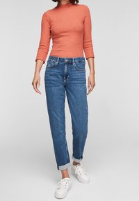 Ein rostfarbener, gerippter Langarm-Top, kombiniert mit Jeans in mittlerem Waschton, leicht zulaufend an den Knöcheln, mit umgeschlagenen Säumen und weißen Sneakers.