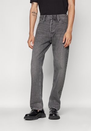 Homme portant un jean noir délavé droit, une chemise noire, des chaussures en cuir noires, debout, les mains détendues le long du corps, sur un fond uni.