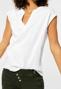 Donna che indossa una blusa senza maniche bianca con un colletto a V e pantaloni verde scuro con tre bottoni visibili.