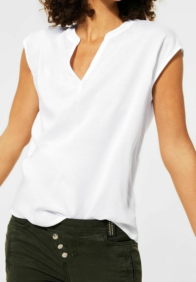 Donna che indossa una blusa senza maniche bianca con un colletto a V e pantaloni verde scuro con tre bottoni visibili.