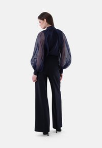 Blusa trasparente blu navy con maniche a sbuffo, collo alto, abbinata a pantaloni neri a gamba larga, silhouette elegante, texture morbida in tutto.