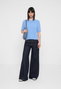 Maglione azzurro chiaro lavorato a maglia con maniche corte, abbinato a jeans larghi di colore blu scuro. L'individuo tiene una piccola borsa blu.