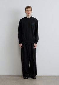 Μαύρο φούτερ με στρογγυλή λαιμόκοψη, με ένα διακριτικό λογότυπο. Συνδυάζεται με φαρδιά μαύρη παντελόνια και μαύρα παπούτσια.