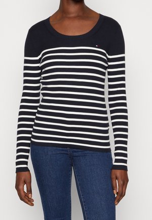 Kvinde iført en marineblå og hvid stribet trøje med lange ærmer, kombineret med blå jeans, stående foran en hvid baggrund.
