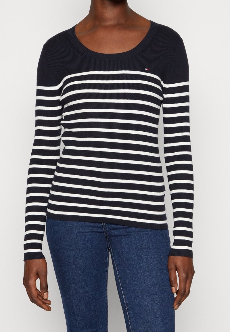 Femme portant un pull à manches longues à rayures horizontales bleu marine et blanc, associé à un jean bleu, debout devant un fond blanc.
