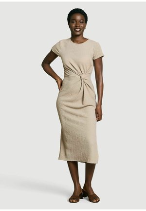 Frau steht mit der Hand an der Hüfte, trägt ein beige kurzärmeliges Midikleid mit vorderem Twist-Detail und braune Sandalen mit offenem Zehenblockabsatz.