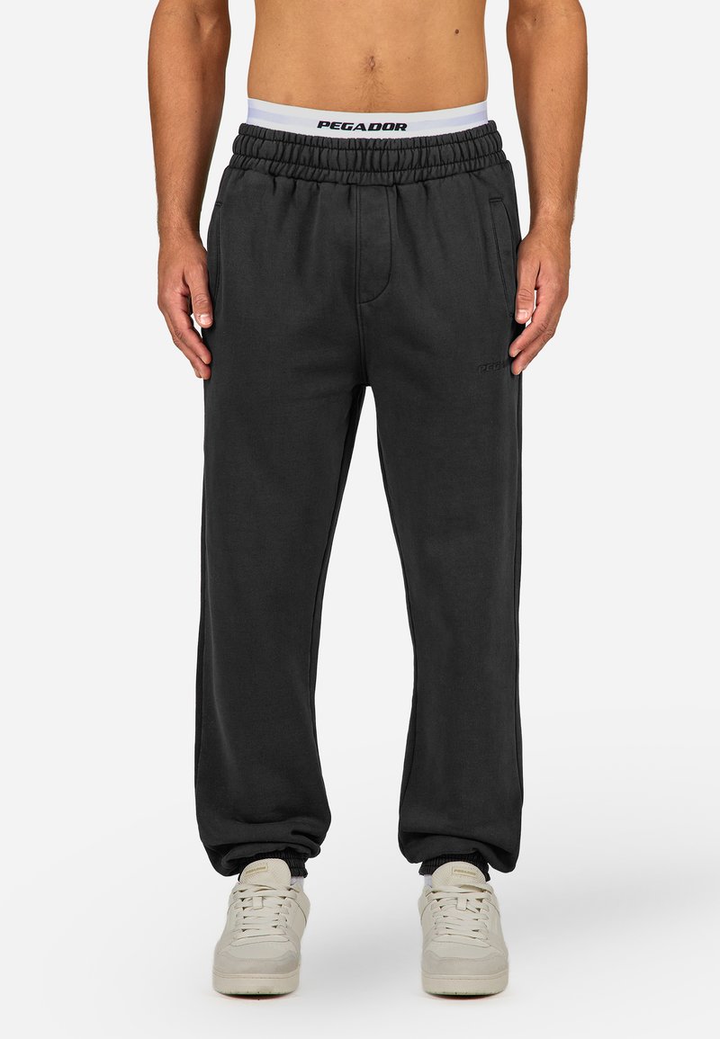 Man wearing black elastic-waist joggers with side pockets and white sneakers, standing against a plain background.