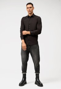 Camicia nera attillata, maniche aderenti, jeans grigio scuro a gamba affusolata e stivaletti neri con suola spessa, su uno sfondo semplice.