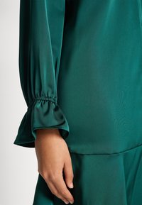 Blusa in satin verde con maniche decorate da volant e polsino arricciato. Texture liscia e lucentezza naturale. Mano appoggiata sul tessuto sotto il polsino.