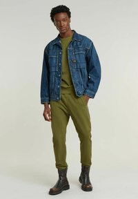 Giacca di denim blu con due tasche sul petto, felpa verde, pantaloni jogger oliva e stivaletti neri con dettagli marroni.