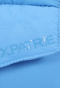 Fénykékkel varrt, sima textúrájú, világoskék párnázott anyag. Fényes, nagybetűs "XPATRIE" logóval és varrott szélekkel rendelkezik.