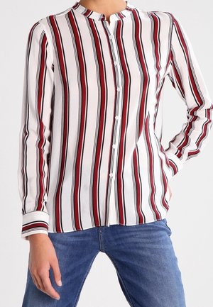 Femme portant une chemise blanche à manches longues avec des rayures verticales rouges et noires, boutonnée à l'avant, associée à un jean bleu, main sur la hanche.