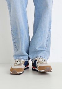 Sneakers with a beige, navy, and brown color scheme. Made of suede and mesh, featuring a textured sole and laces, paired with light blue jeans.