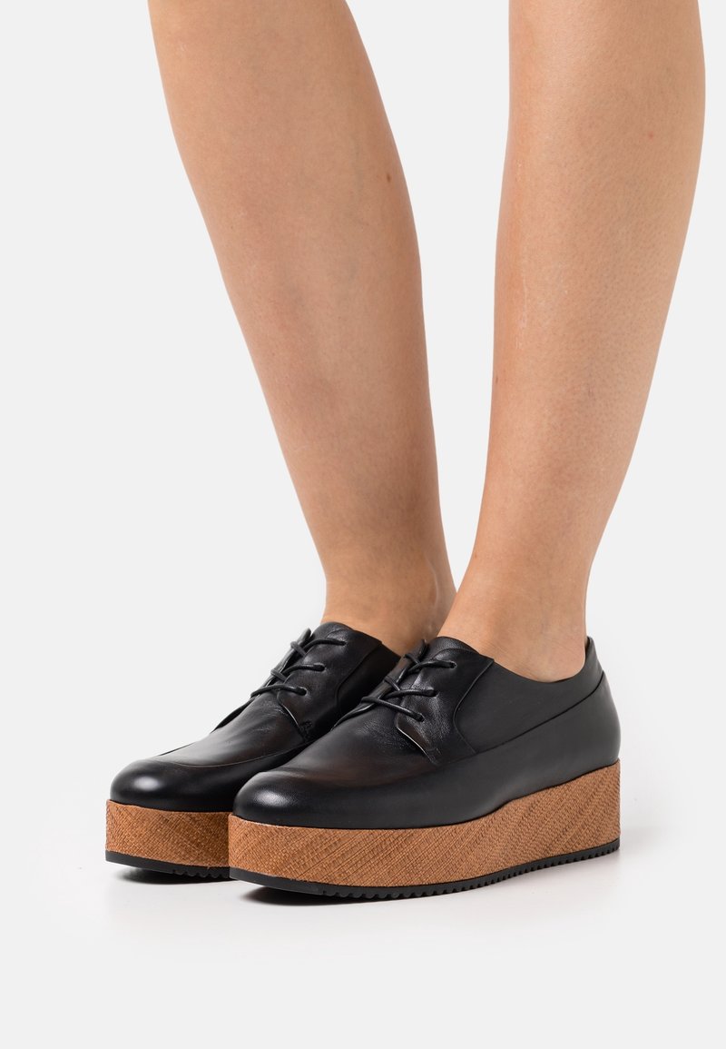 Chaussures à plateforme en cuir noir à lacets avec semelles épaisses marron texturées portées sur des jambes nues sur fond blanc uni.