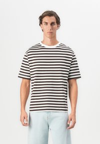 Mann som har på seg en svart og hvit horisontalt stripete t-skjorte og lyseblå jeans, stående mot en ensfarget hvit bakgrunn.