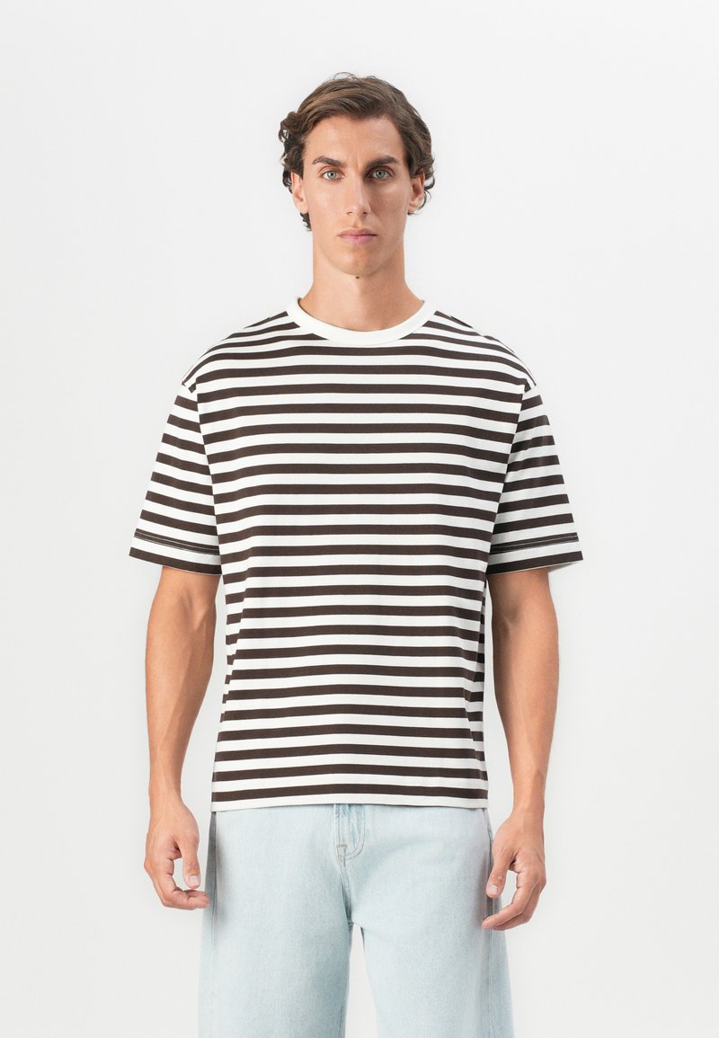 Mann som har på seg en svart og hvit horisontalt stripete t-skjorte og lyseblå jeans, stående mot en ensfarget hvit bakgrunn.