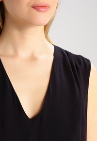 Svart ärmlös topp med en djup V-ringning, tillverkad av ett slätt material; har mjuka veck nära axlarna.