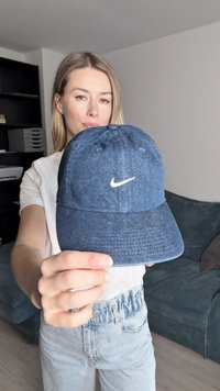 Temno modra jeans kapica za bejzbol z belim logotipom Nike na sprednji strani. Ima šivane panele in nastavljiv pas na hrbtni strani.