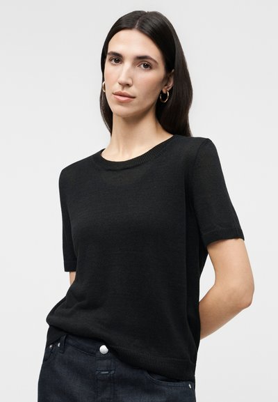 Γυναίκα με μακριά σκούρα μαλλιά που φορά χρυσούς κρίκους σκουλαρίκια, μαύρη κοντομάνικη πλεκτή μπλούζα και σκούρα τζιν τζιν παντελόνια, μπροστά σε απλό φωτεινό φόντο.