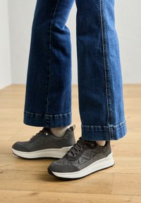 Zapatillas gris texturizadas con una suela blanca, que presentan un patrón geométrico y un diseño de cordones, combinadas con jeans de pierna ancha azules.