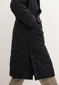 Cappotto lungo trapuntato nero con vita regolabile e bottoni a pressione, caratterizzato da un motivo testurizzato e una fessura laterale per facilitare i movimenti.