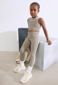 Ensemble de sport beige comprenant un crop top ajusté et des leggings taille haute avec des bandes blanches sur le côté. Accompagné de baskets légères et de chaussettes blanches.