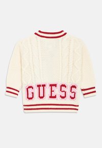 Maglione lavorato a maglia color crema con motivo a trecce, bordatura a righe rosse e grande logo "GUESS" in rosa e rosso sulla parte bassa della schiena. Maniche corte.