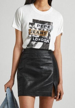 Weißes grafisches T-Shirt mit paillettenbesetztem Schriftzug, kombiniert mit einem schwarzen Minirock aus Leder mit seitlichem Schlitz. Die Jeansjacke wird in einer Hand gehalten.