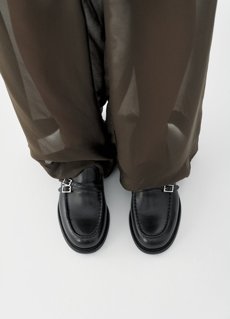 Μαύρα δερμάτινα παπούτσια με στρογγυλεμένη μύτη, που διαθέτουν δύο αγκράφες και υφή στη σόλα, συνδυασμένα με ημιδιάφανο σκούρο καφέ παντελόνι.