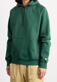 Vihreä huppari, valmistettu paksusta kankaasta, jossa kengurun tasku, kiristettävä huppu, resorit hihansuissa ja pieni logo hihassa.