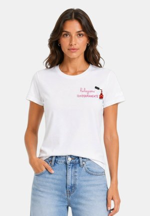 Donna con capelli castani che indossa una maglietta bianca con testo rosa e un motivo di smalto per unghie, abbinata a jeans azzurri.