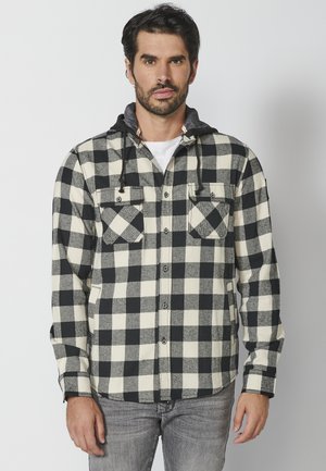 Camisa de franela a cuadros en blanco y negro con capucha gris, que cuenta con dos bolsillos en el pecho y un cierre frontal de botones. Textura suave, diseño casual.