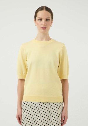 Vrouw met lichtgeel kort mouwtruitje en witte rok met zwarte stippen, staand tegen een effen lichte achtergrond.